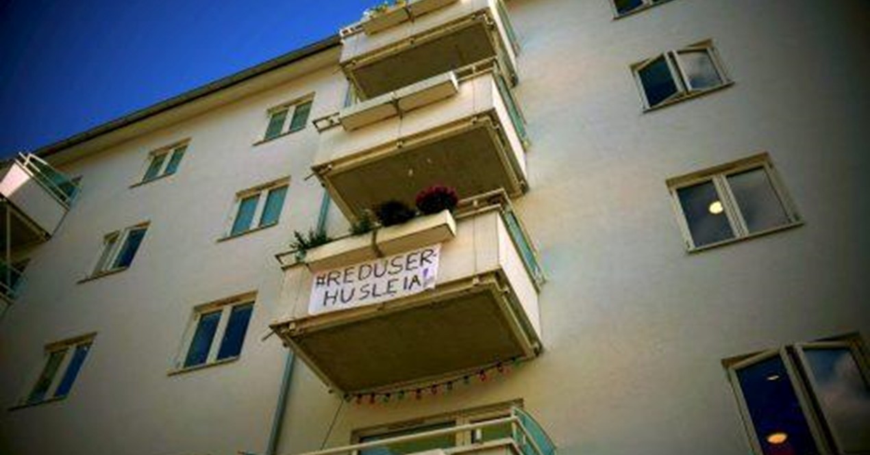 Bilde av bygård med et banner hengende fra balkongen, det det står "Reduser husleia"