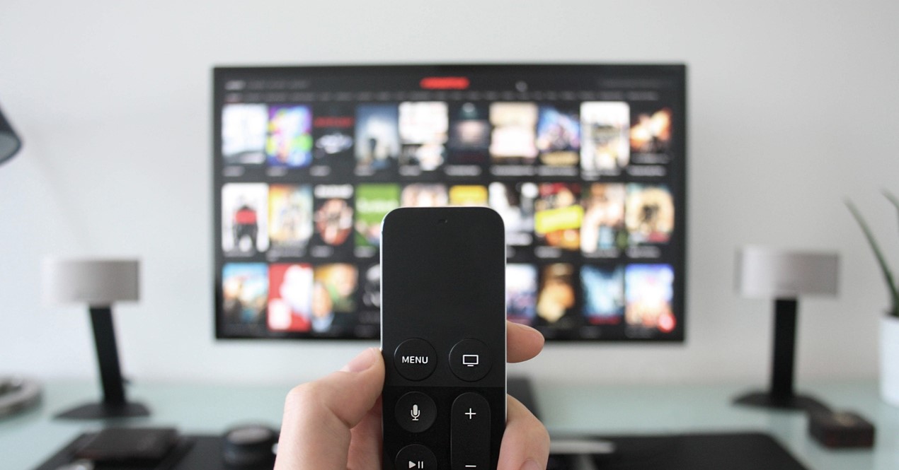 Fjernkontroll i en hånd i fokus med en påslått TV i bakgrunnen